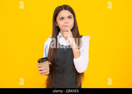 Thinking pensive clever teenager girl. Teenager child with coffee cup isolated on yellow studio background. Girl drinking take away beverage. Stock Photo