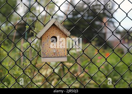 Typical common wooden birdhouse hanging on a chain link fence in the sunny summer meadow. Stock Photo