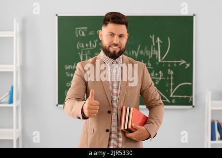 Handsome Math teacher with books showing thumb-up in classroom Stock Photo