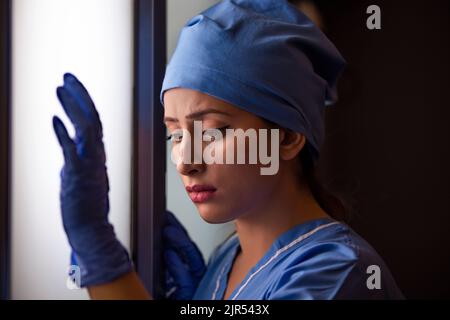 Close-up of thoughtful young female nurse standing by window in hospital. Stock Photo