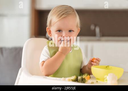 Happy blond baby in green bib eating with pleasure organic steamed carrot and broccoli by hand, sitting in high chair at table with plate on kitchen background. Baby-led weaning. Self-feeding concept Stock Photo