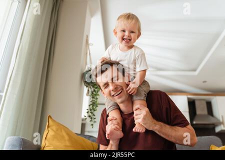 Laughing father giving his kid piggyback ride on shoulders, baby boy pulling daddy's hair, father and son spending time together on sunday morning in living-room. Dad playing with his toddler boy Stock Photo