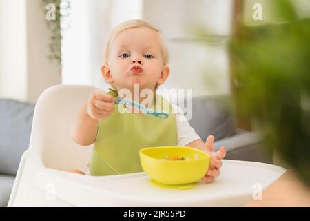 Adorable blond baby boy sitting at table in high chair with spoon in hand and yellow plate with breakfast in front of him, training skills to feed himself, wearing green bib. Baby eating by himself Stock Photo