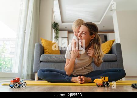 Funny picture of cheerful mother sitting in butterfly position on mat practicing yoga piggybacking her cute baby boy in living-room with gray couch on background. Healthy motherhood and wellness Stock Photo