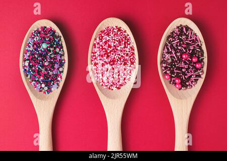 Colourful cake sprinkles in wooden spoons on red paper background. Baking decorations, top view. Stock Photo