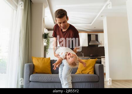 Young father spending time with his baby boy, cuddling and playing active games in living-room after returning home from work, carrying child as airplane, swaying together on background with gray sofa Stock Photo