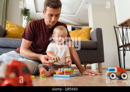 Father assisting his baby boy playing sorter sitting on floor in living-room with sofa on background, surrounded with car toys, waiting mom to come home from work. Education toys, child development Stock Photo