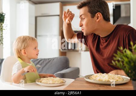 Happy dad playing with spaghetti while having dinner with his baby boy in bib learning to eat with fork looking at funny dad with interest. Happy fatherhood, feeding kid. Father day, family Stock Photo
