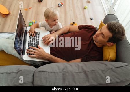 Naughty little baby boy bothering his father lying on couch with laptop in living-room, tapping keyboard with hand, disturbing working from home or resting dad. Toddler son asking for dad attention Stock Photo