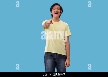 Portrait of a teenage boy pointing towards camera while standing against blue background Stock Photo