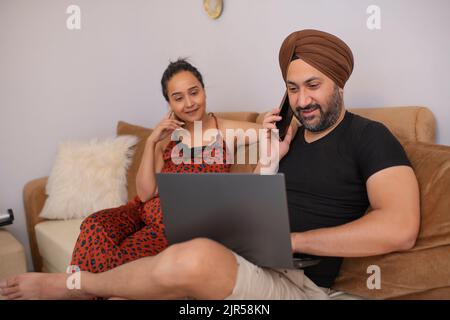 Sikh man talking on Smartphone while using laptop and woman sitting beside Stock Photo