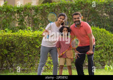 Happy parent with their child posing in front of camera with holding badminton racquets in the park Stock Photo