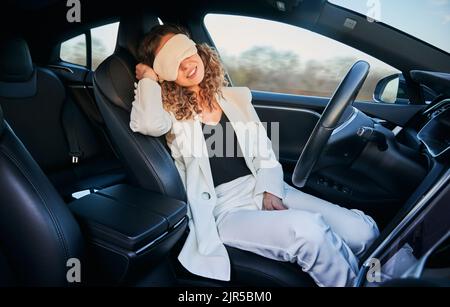 Charming woman in sleeping mask sitting on driver seat and smiling while taking nap in electric car with autopilot system. Businesswoman using driver-assistance system while sleeping in automobile. Stock Photo