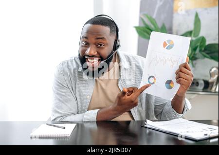 Clever positive african american man with headset, business leader, financial mentor, having online business conversation, records online lesson, conducts consultation, shows financial graph, smiles Stock Photo