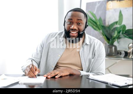 Confident african american man with headphones, business leader, company employee, having online business conversation, listens to the interlocutor, conducts consultation, takes notes, smiles Stock Photo