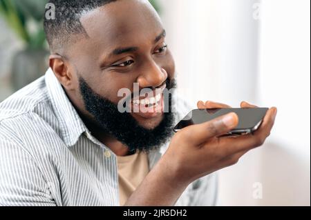 Recording a voice message. Wireless technology. Close up of a positive african american man holding his smartphone near his face, talking on speakerphone, recording audio message, smiling Stock Photo