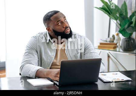 Pensive upset African American businessman, ceo, office worker, sits in modern office at a table, at his workplace, looks thoughtfully to the side, thinks about a new project, dreams of rest, vacation Stock Photo