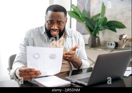 Confused puzzled african american business man, freelancer or company ceo, working with documents, sit at desk in modern office, working on a project uses laptop, studying financial graphs, stunned Stock Photo