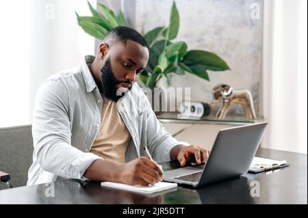 Focused successful smart african american male company owner, manager, sitting at a desk in modern office, working on a project, with documents, analyzing, taking notes, prepares a report Stock Photo