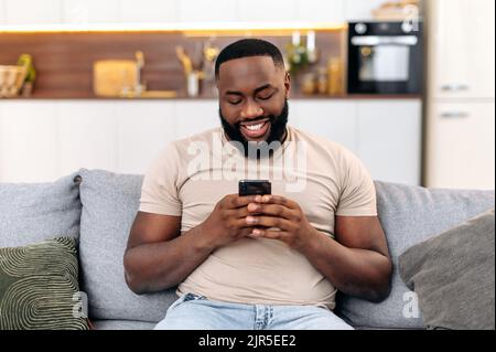 Modern attractive positive african american man in casual wear, sits at home on sofa, using his smartphone, texting online, browsing social networks, messaging with friends, answers email, smiles Stock Photo