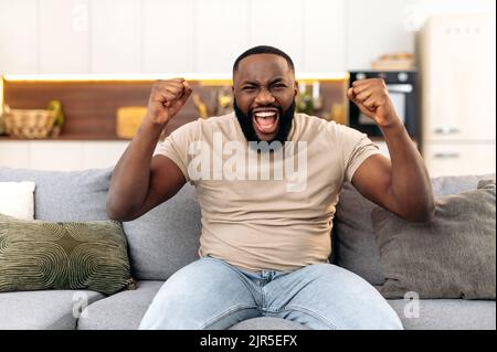 Winning concept. Amazed cheerful young african american man, sitting on the sofa in living room, excited for success with arms raised, celebrating victory, rejoices at a goal, smile happily, screaming Stock Photo