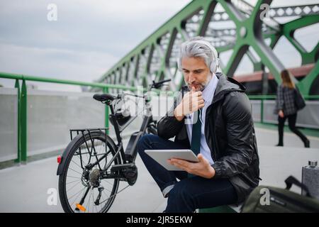 Businessman with bike sitting on bridge, using tablet and listening to music. Commuting and alternative transport concept Stock Photo