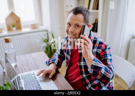 Young man with Down syndrome sitting at desk in office and using smartphone and laptop, looking at camera and smiling. Concept of integration disabled Stock Photo