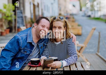 Portrait of happy young man with Down syndrome with his mother sitting at cafe outdoors and talking. Stock Photo