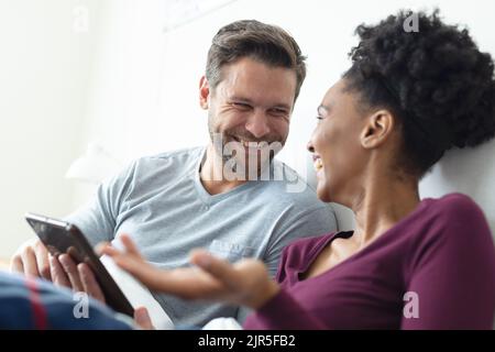 Image of happy diverse couple talking in bed and using tablet Stock Photo