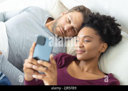 Image of diverse couple using smartphone and lying in bed Stock Photo