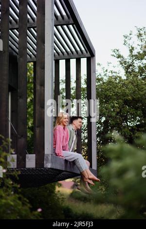 Young couple sitting and cuddling in hammock terrace in their new home in tiny house in woods, sustainable living concept. Stock Photo