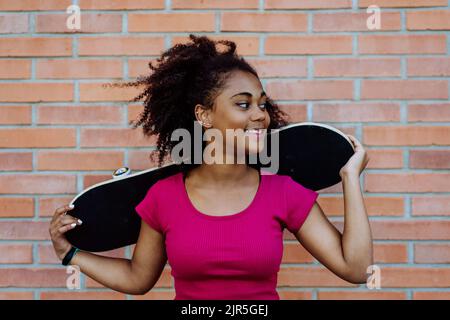 Multiracial teenage girl with backpack and skateboard, walking in city during summer day. Stock Photo