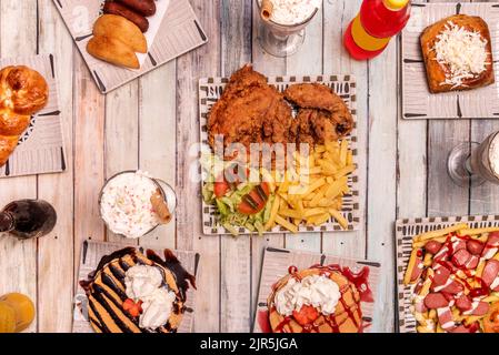 Set of fast food dishes with chicken in batter with potatoes and salad, pancakes with chocolate and strawberry syrup, lots of cream and dumplings Stock Photo