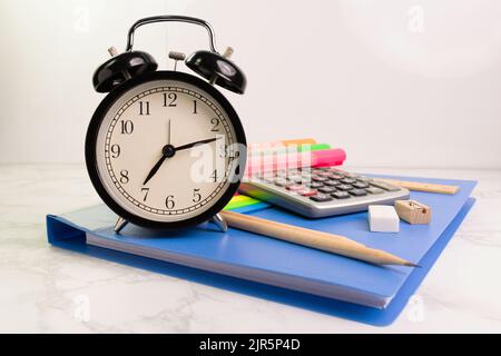 black clock with 7 am 12 minutes with blue folder and writing equipment with Calculator white table at the office Stock Photo
