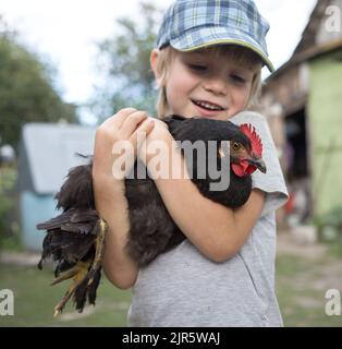 Caucasian boy holding a chicken in his arms. Happy child spends summer on the farm. Treating poultry, interesting childhood. Selective focus Stock Photo