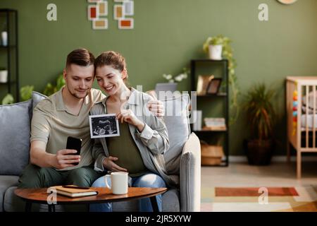 Young couple sitting on sofa in living room and showing ultrasound image during video call on mobile phone Stock Photo