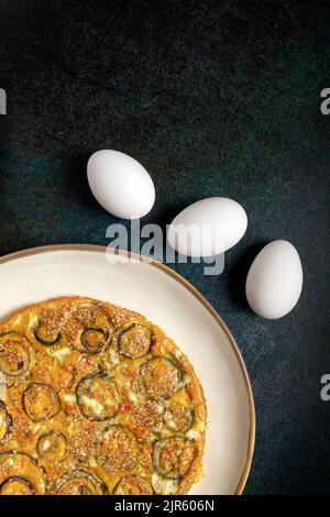 Homemade onion omelette in a white dish beside three fresh eggs on a raw dark background. Stock Photo