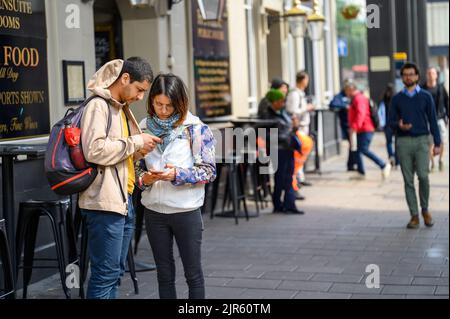 LONDON - May 20, 2022: Man and woman with backpacks looking at smartphones on busy London street Stock Photo