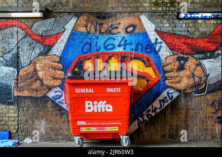 LONDON - May 20, 2022: Large red wheeled bin in front of NHS super hero graffiti on old brick wall Stock Photo