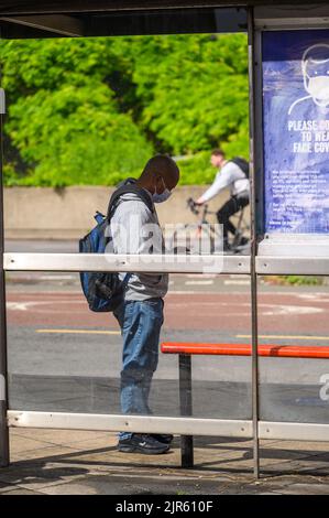 LONDON - May 20, 2022: Man wearing face mask looks at smartphone while waiting at bus stop Stock Photo