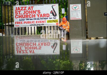 LONDON - May 20, 2022: Construction worker wearing orange hard hat and reflective jacket sits using smartphone Stock Photo