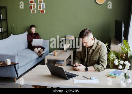 Young man sitting at his workplace with laptop and using mobile phone for work with his girlfriend working in background Stock Photo