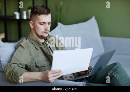 Young serious man sitting on sofa with laptop on his knees,and typing document online during remote work at home Stock Photo