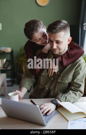 Young smiling woman embracing her boyfriend while he doing his online work on laptop at home Stock Photo