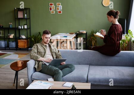 Young man working online on laptop while sitting on sofa with woman reading book and drinking coffee sitting opposite him Stock Photo