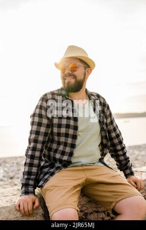 Handsome and confident. Outdoor portrait of smiling man wearing hat and sunglasses on beach. Holidays travel and summer tourism Stock Photo