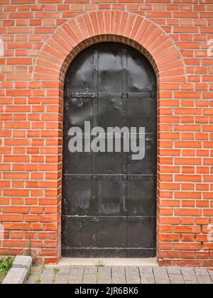 Old metal arch type door in the red brick wall. Black metal grunge gate. Stock Photo