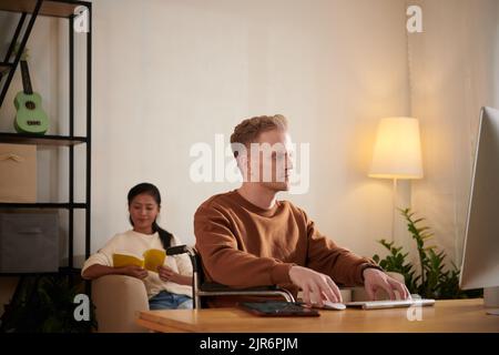Serious young man in wheelchair working on computer, his girlfriend reading book in background Stock Photo