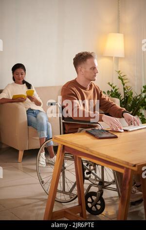 Serious young man in wheelchair working on computer at home, his wife reading in background Stock Photo
