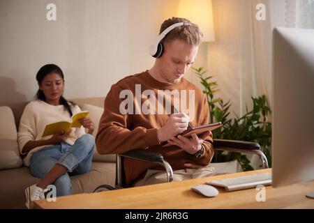 Young man with disability listening to music and working on tablet computer at home, girlfriend reading book in background Stock Photo